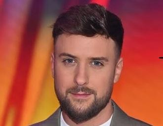 Aleksander Sikora, prezenter telewizyjny, uśmiechnięty i gestykulujący w nowoczesnym studiu telewizyjnym.