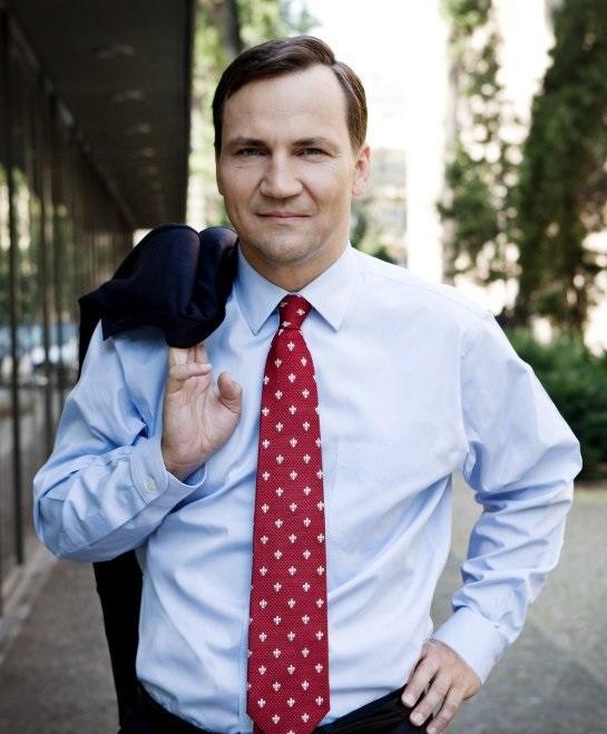 Radosław Sikorski podczas spotkania dyplomatycznego, aktywnie gestykulujący, w tle zamazane postaci i godło Polski, symbolizujące jego rolę w polityce zagranicznej.