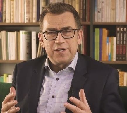 Maciej Orłoś, dziennikarz i autor, w swobodnej pozie, gestykuluje, na tle rozmytej biblioteczki, symbolizującej jego działalność literacką i szkoleniową.