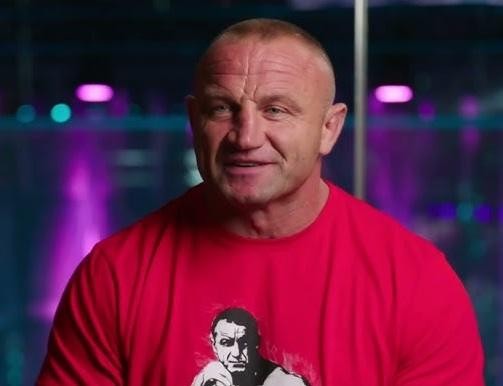 Mariusz Pudzianowski prezentujący siłę i muskularną sylwetkę, na tle symbolizującym jego karierę strongmana i zawodnika MMA.